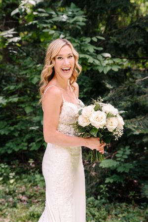 A bride in a white lace gown holds a cream rose bouquet against a garden backdrop.