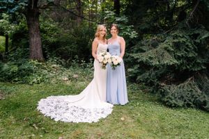 A bride in lace gown with long train poses with attendant in light blue dress in a garden setting.
