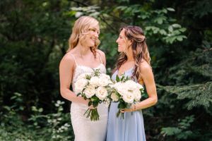 Two women in formal dresses share a joyful moment while holding white floral bouquets in a forest setting.