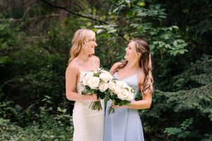 Two women in elegant dresses share laughter while holding white bouquets in a woodland setting.