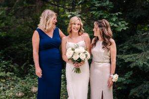 Three women in elegant dresses pose together while holding beautiful white bouquets against greenery.