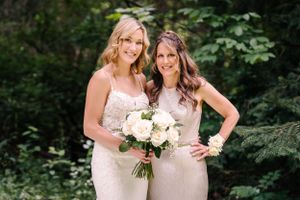 Two women in formal attire pose together with white bouquets against a lush forest background.