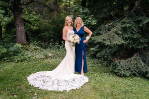 White wedding gown with long train contrasts with navy blue dress as two women pose in garden setting.