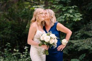Two women in contrasting formal dresses share a candid moment with white bouquet in forest setting.