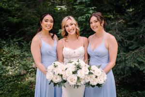 Three women in pale blue and white dresses hold matching white floral bouquets against forest backdrop.