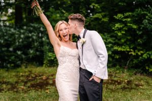 Joyful wedding couple celebrating with raised bouquet in outdoor forest setting.