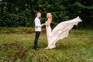 Wedding dress billowing dramatically in the wind as couple poses in forest clearing.