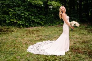 Bride in flowing lace gown holds bouquet while posing in outdoor garden setting.