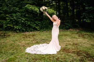 Bride raises bouquet triumphantly while wearing fitted lace wedding gown outdoors.