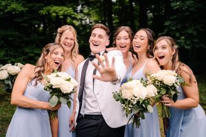 Joyful wedding party laughs together outdoors with bridesmaids holding white bouquets.