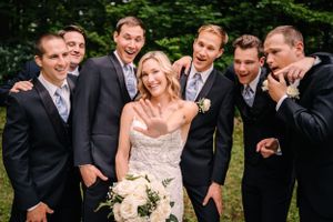 Wedding party poses playfully in outdoor garden setting with groomsmen in dark suits and bride in lace gown.