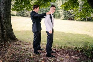 Wedding day preparations as groomsman helps adjust groom's attire under tree.