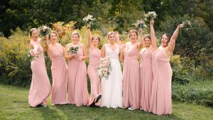 Bridal party in pink dresses celebrates joyfully in garden setting.