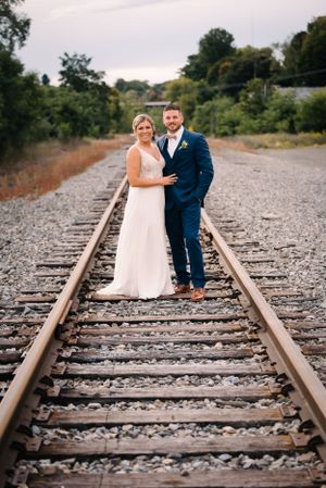 Wedding couple stands centered on converging train tracks during outdoor photoshoot.