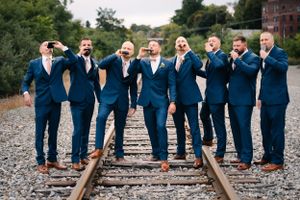 Groomsmen strike casual poses while standing on railroad tracks in navy suits.
