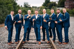 Groomsmen in matching navy suits pose confidently on railroad tracks with red brick building behind.