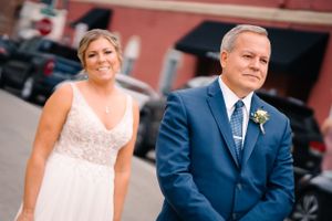 Wedding day portrait outdoors with navy suit and white lace bridal gown against brick building.