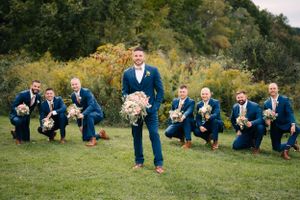 Wedding party groomsmen pose playfully on grass with navy suits and brown shoes against autumn foliage.