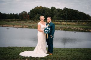 A couple poses in formal wedding attire by a serene lake with trees in the background.