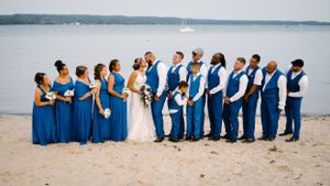 Wedding party in blue attire poses on beach with water and sailboats in background.