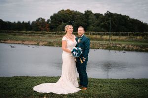 Wedding couple stands together on grass near water with beautiful natural backdrop.