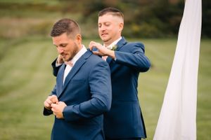 Two people in matching navy suits help each other get dressed for a wedding ceremony.