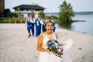 A radiant bride holding vibrant blue bouquet laughs on beach with wedding party behind her.