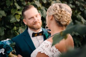Close-up of wedding couple sharing tender moment among green foliage with blue floral bouquet.