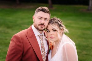 Elegant outdoor wedding portrait with a rust-colored suit and intricate bridal hairstyle.