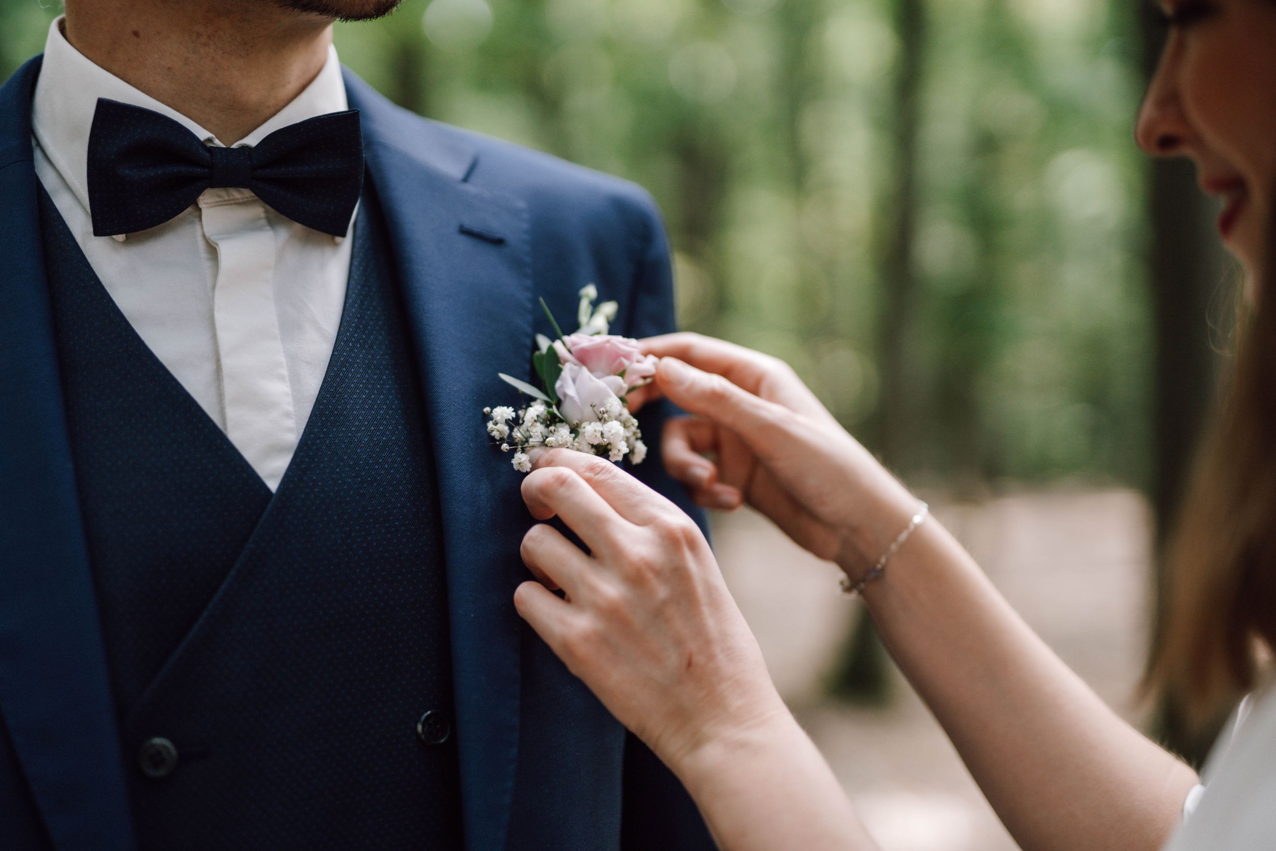 Hands attaching a delicate floral boutonniere to a navy blue suit lapel.