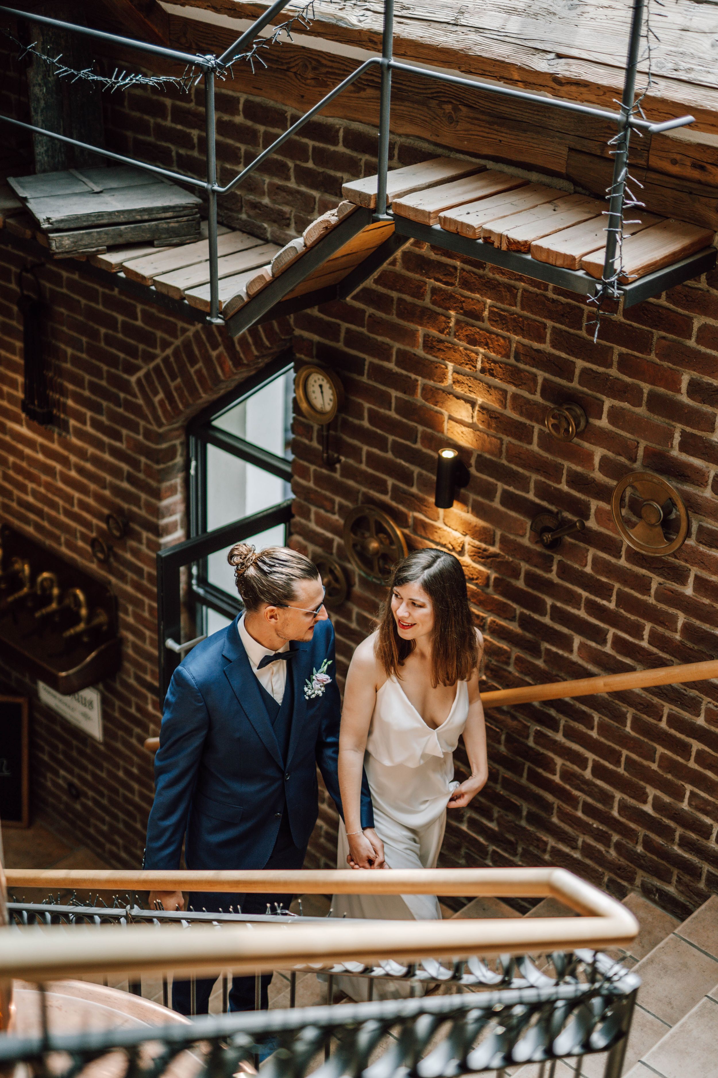 Couple in wedding attire standing in an industrial brick stairwell with warm lighting.