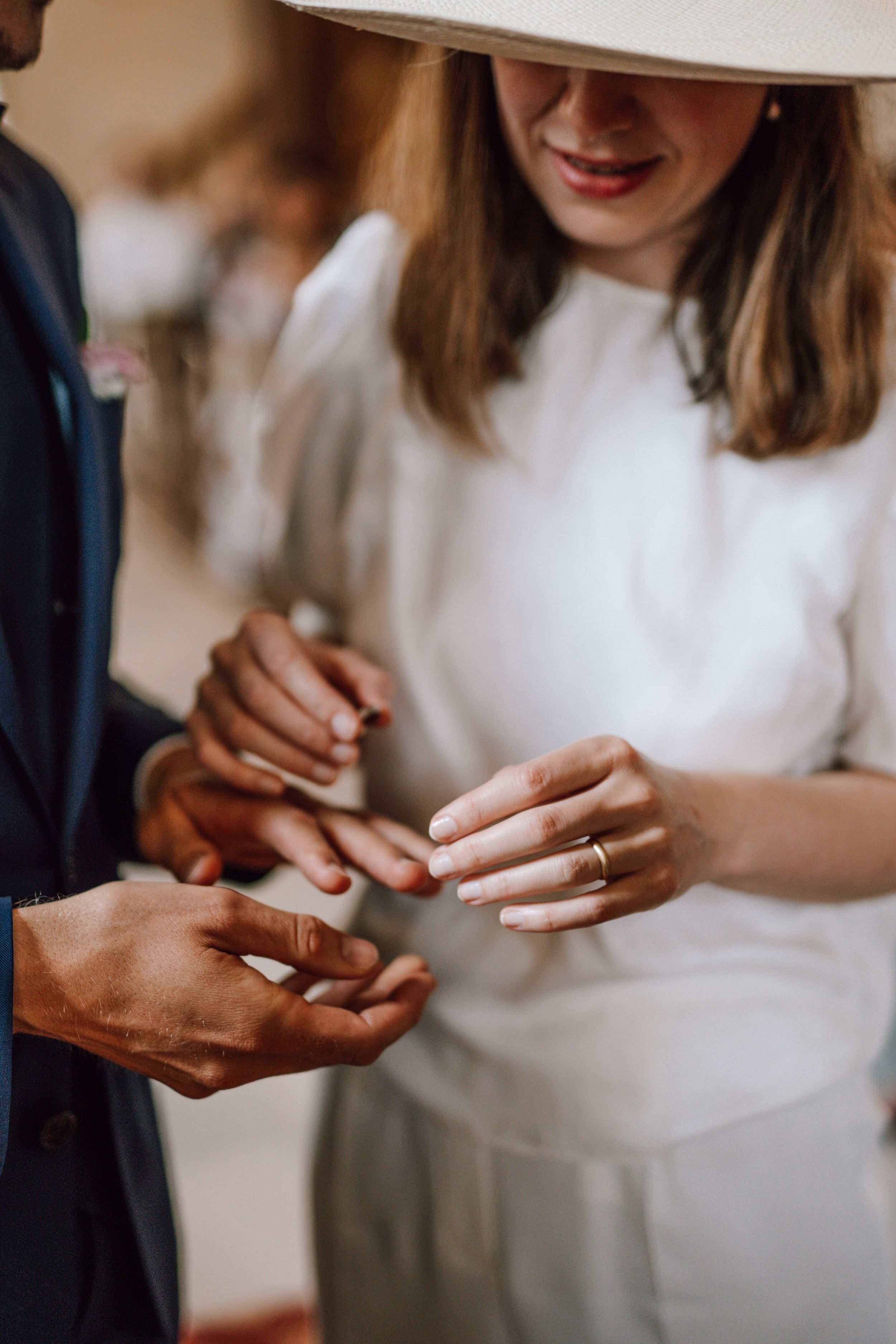 Hands exchanging wedding rings during ceremony, with white dress and navy suit visible.