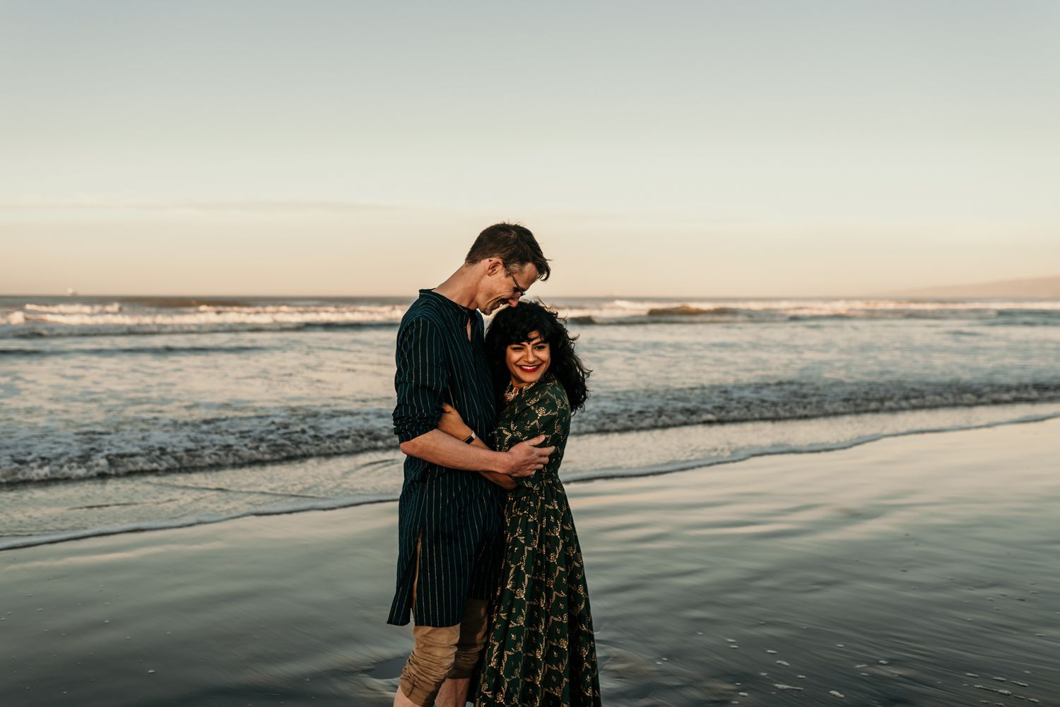 A romantic couple embraces on a beach at sunset with waves crashing in the background.
