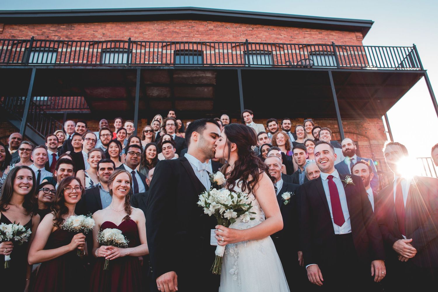 Large wedding party gathered on rustic building balcony at sunset.