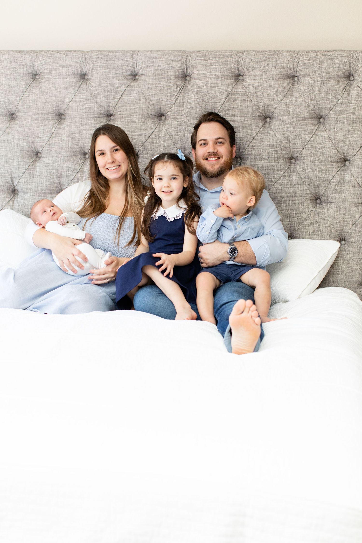 Family lounging together on bed with gray tufted headboard in bright room.