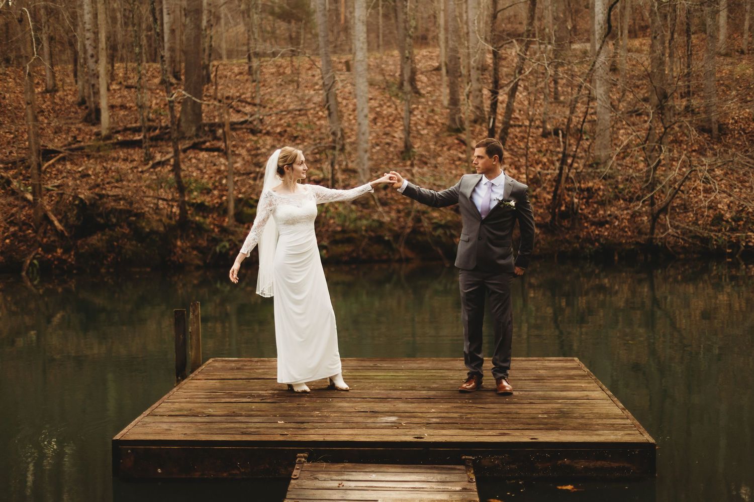 Couple in wedding attire holds hands while standing on wooden dock over autumn lake.