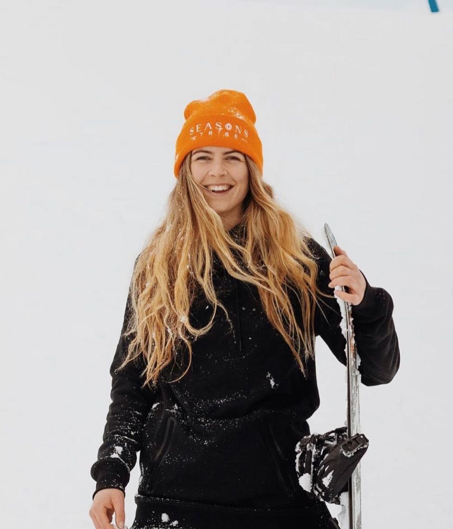 Winter sports enthusiast in orange beanie and black jacket holds ski poles in snowy setting.