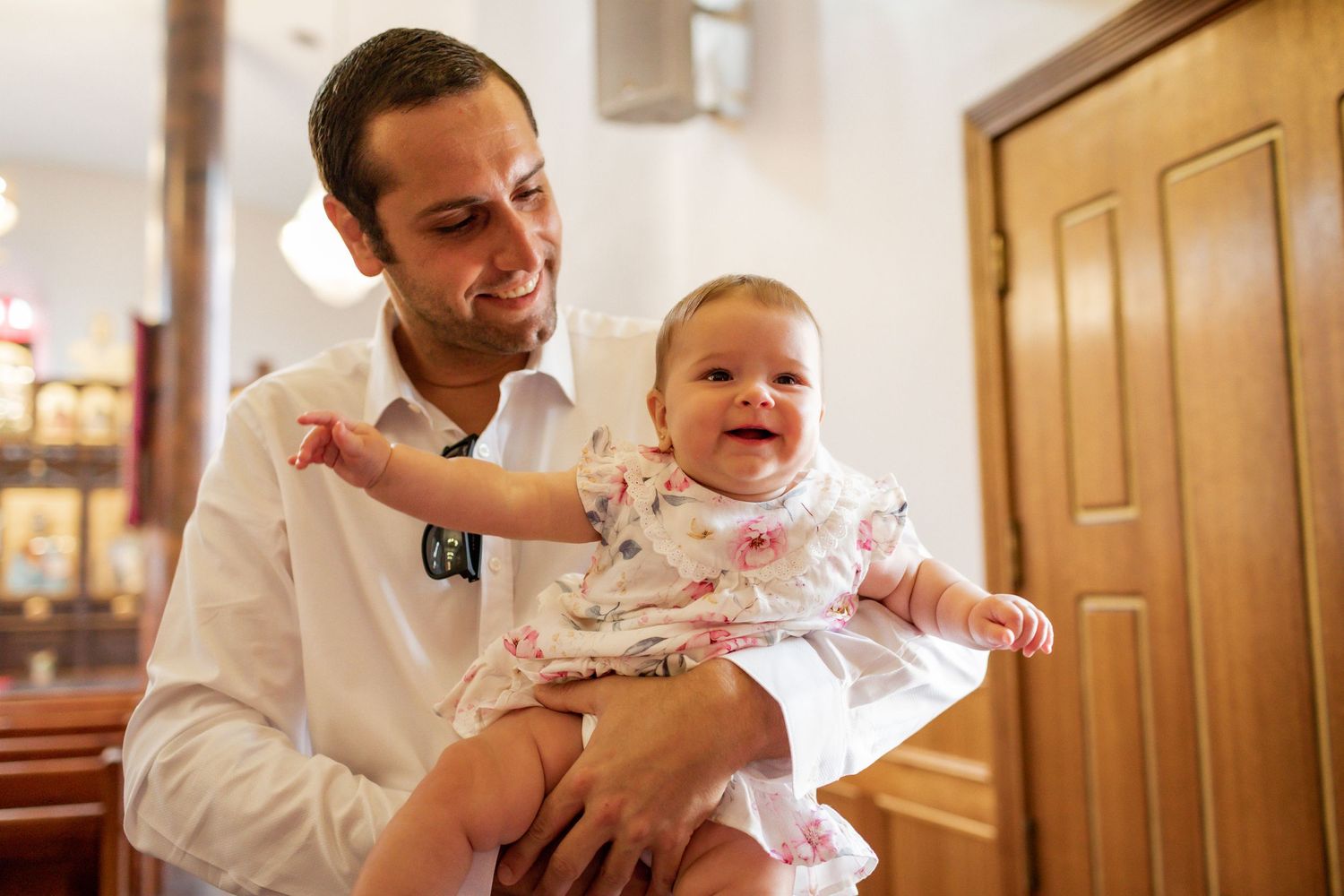 A joyful moment between parent and baby in formal white clothing inside a church.