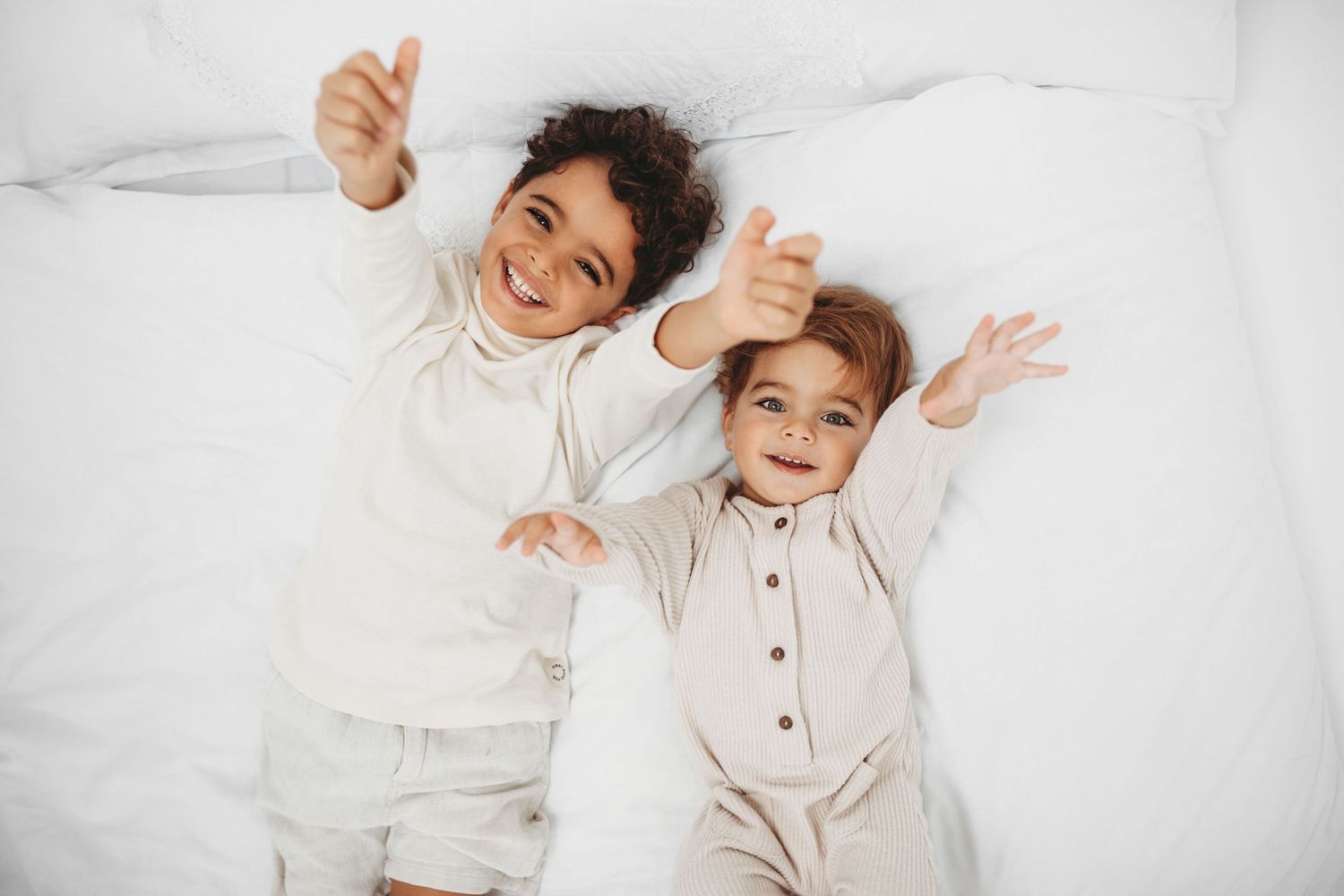 Two children in light colored pajamas lying on white bedding with arms raised joyfully.