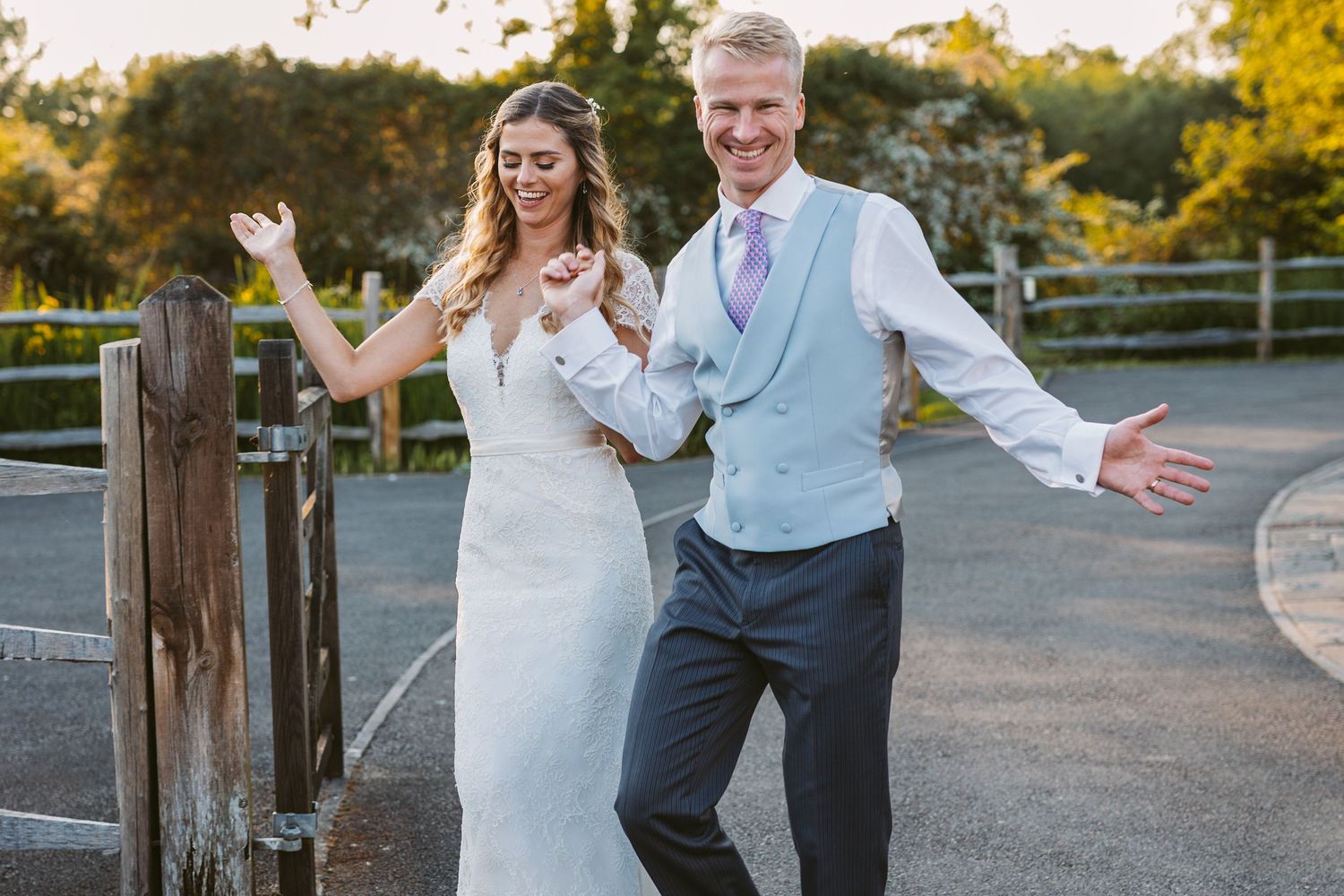 Newlyweds posing playfully by a rustic wooden fence at sunset.