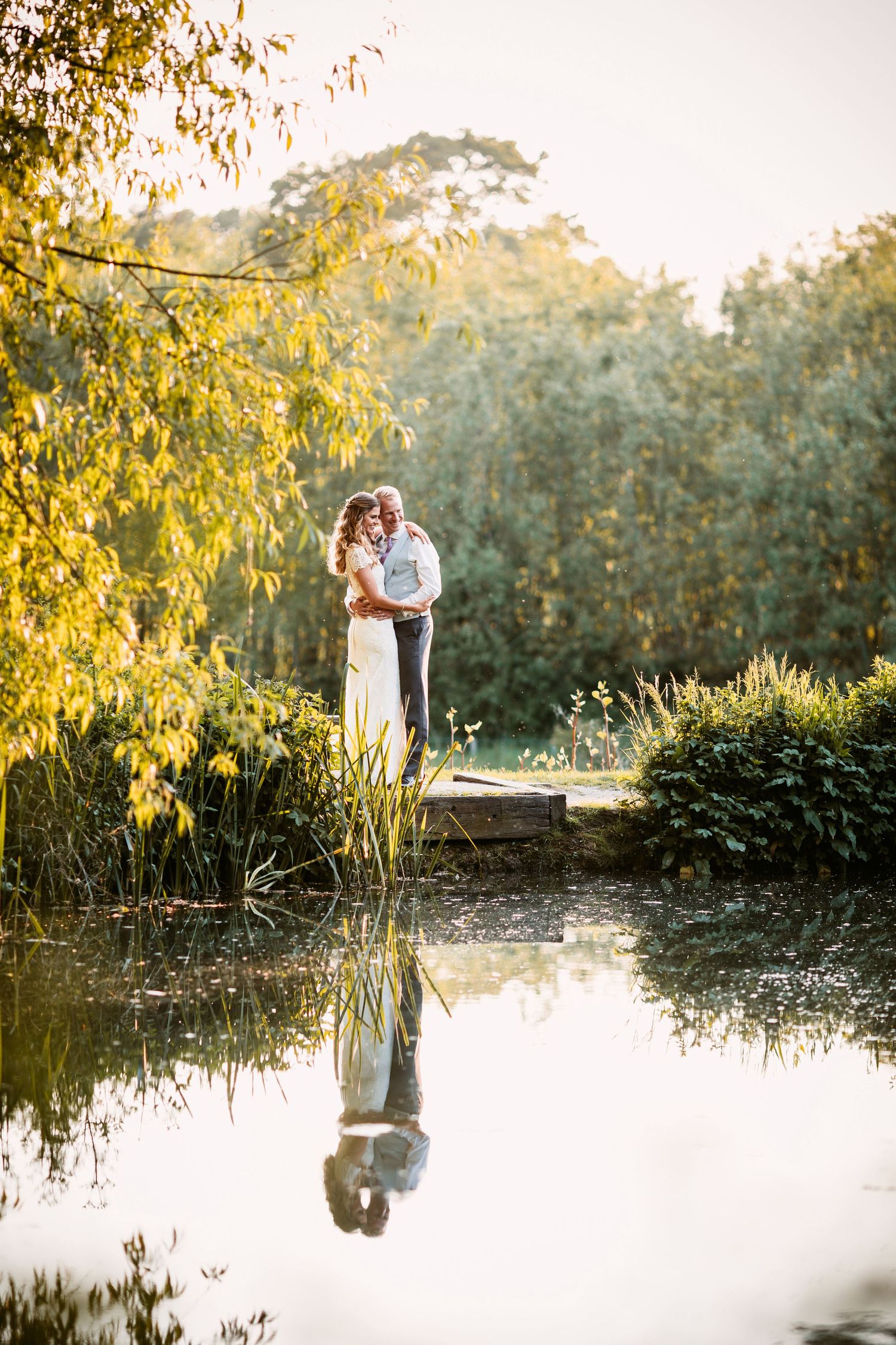 Couple embracing near a pond with their reflection in the water at sunset.