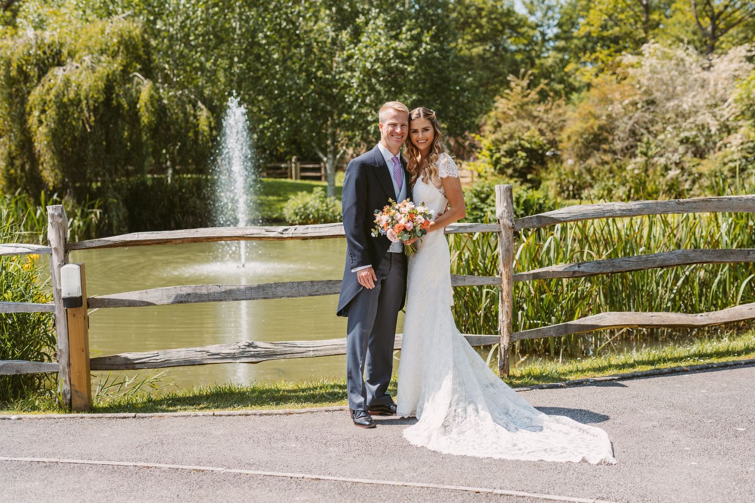 A couple poses for wedding photos by a scenic pond with a fountain in a landscaped garden.