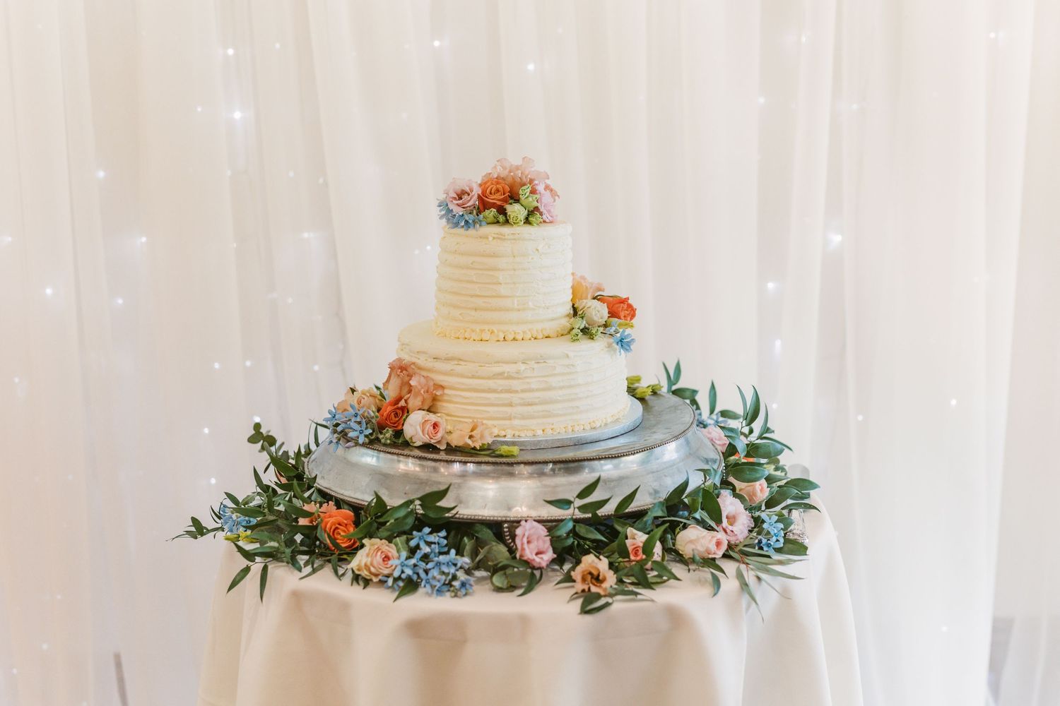 Elegant white wedding cake decorated with fresh flowers and greenery on a draped table.