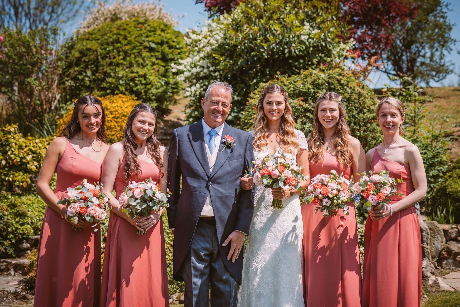 Wedding party poses outdoors with bridesmaids in coral dresses holding floral bouquets.