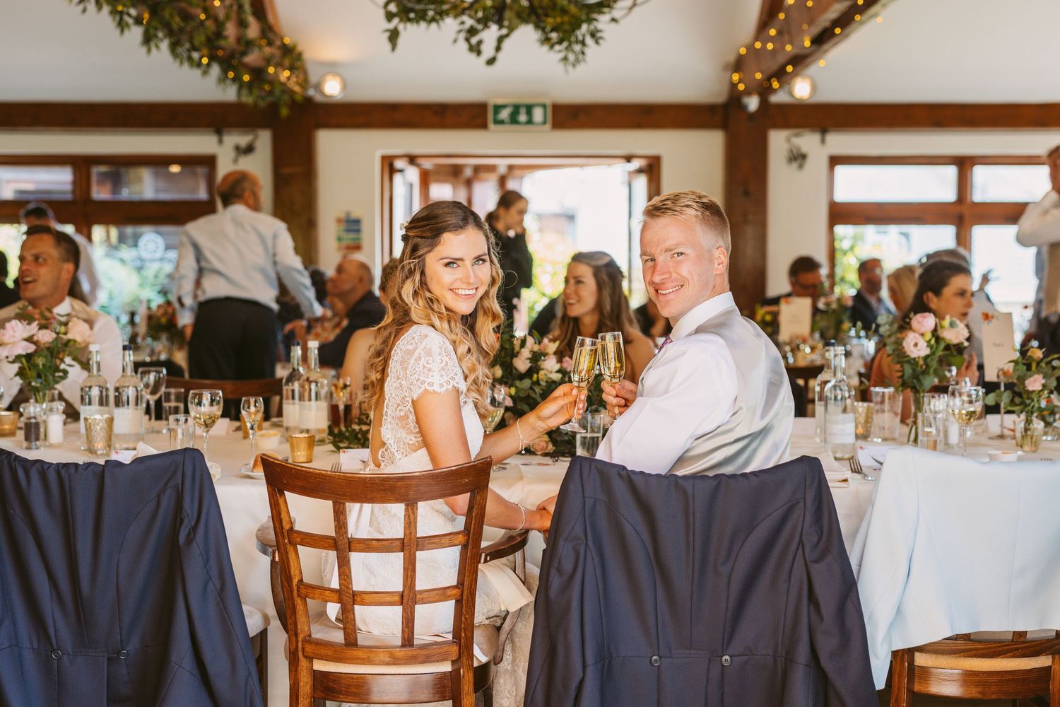 Wedding reception interior with decorated tables and greenery hanging from wooden beams.