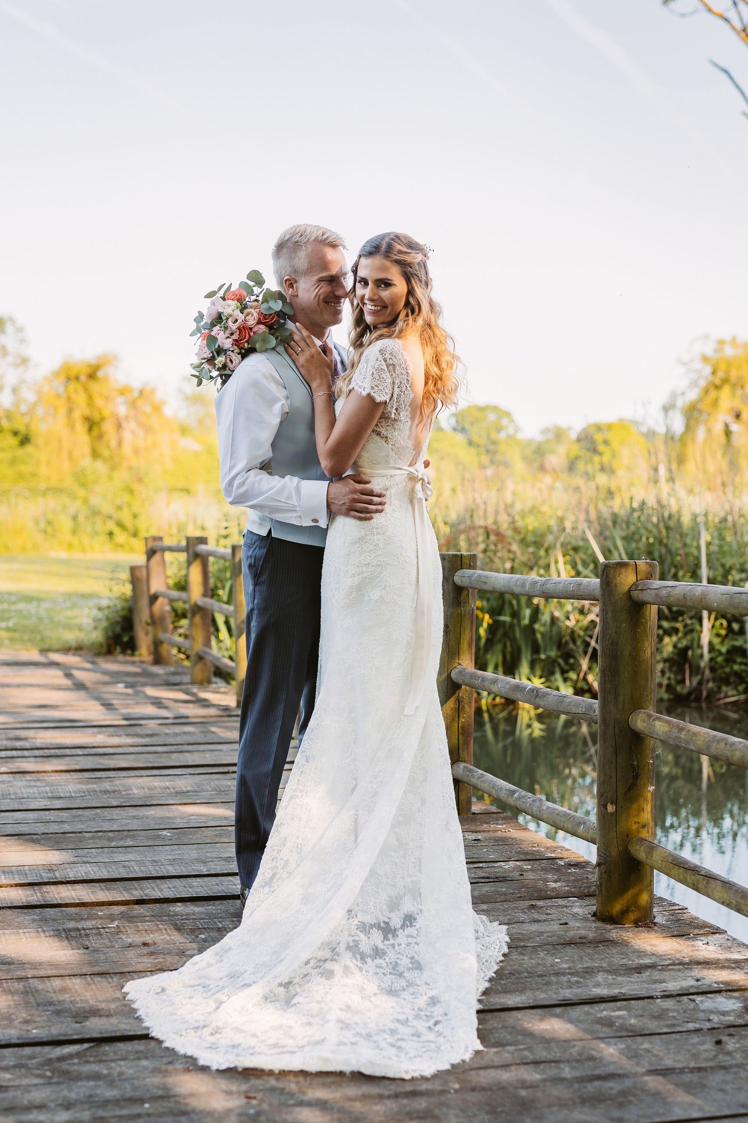 A romantic wedding couple embraces on a rustic wooden bridge surrounded by lush greenery during golden hour.