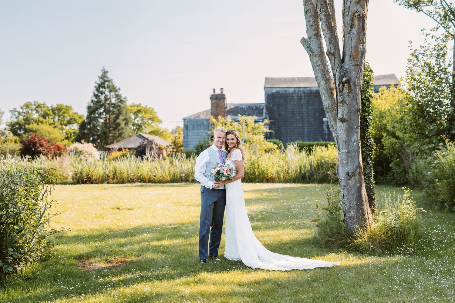 A wedding couple poses in front of a historic building surrounded by lush summer gardens.