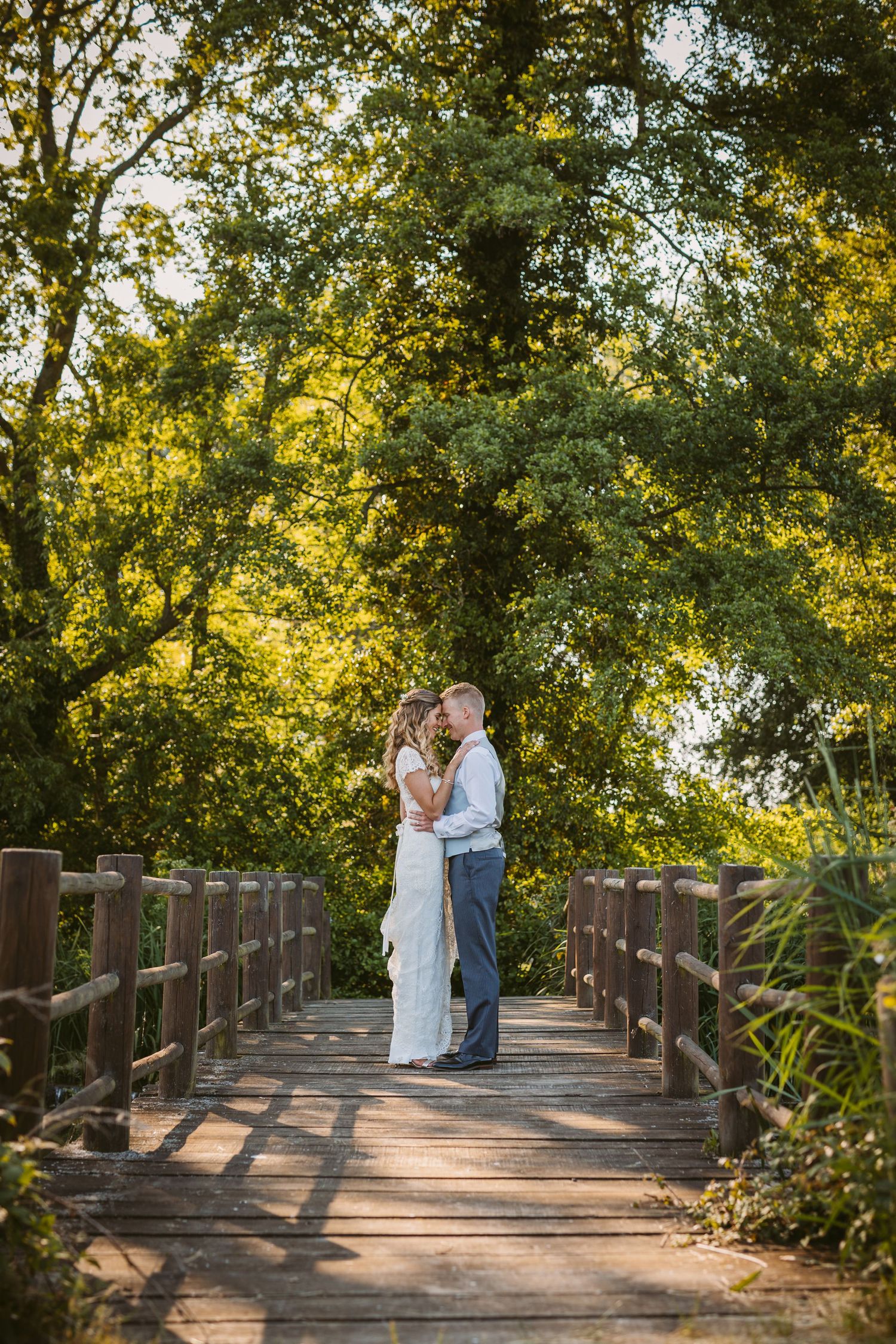 A romantic wedding photo on a wooden bridge surrounded by sunlit trees and nature.