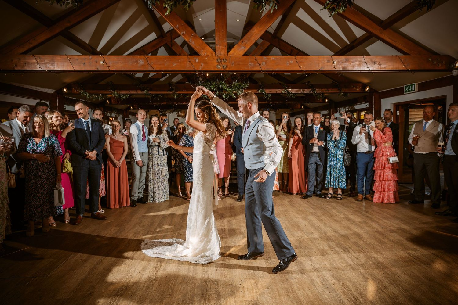A couple shares their first dance surrounded by wedding guests in a rustic wooden venue with exposed beams.