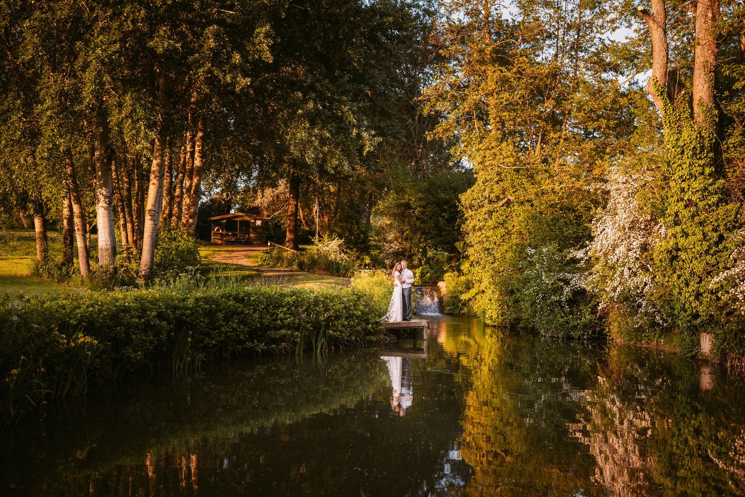 A romantic sunset wedding photo by a reflective pond surrounded by trees.